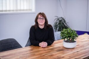 A woman sitting at a wooden table with her hands clasped, smiling at the camera, with a potted plant to her right and an office background with a window and a second potted plant.