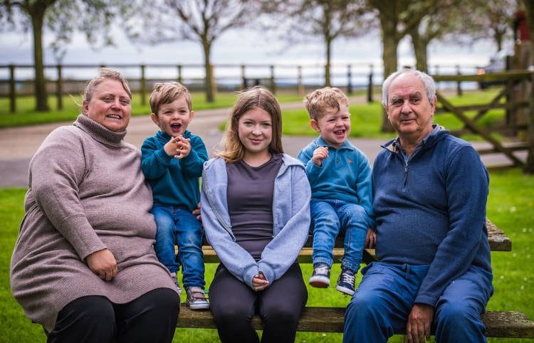 A family of five with two grandparents sitting on a park bench; from left to right, there's an elderly woman, two young boys, a teenage girl, and an elderly man. The children are playful, and everyone is looking towards the camera with a park visible in the background.