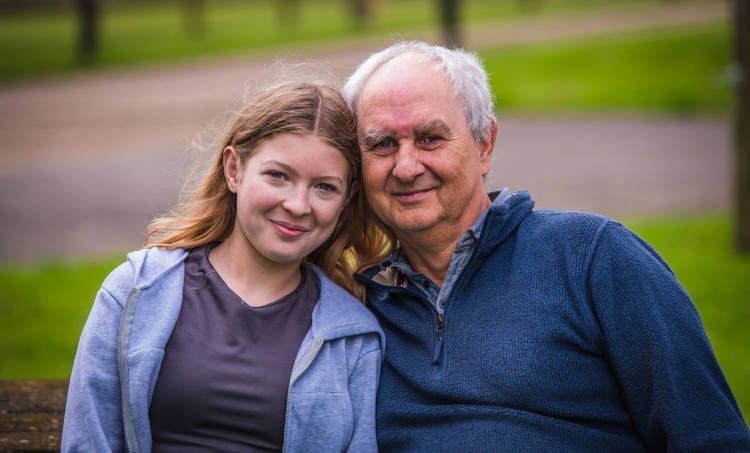 A young woman and an older man smiling at the camera while sitting closely together on a park bench, with a blurred natural background suggesting a peaceful outdoor setting.