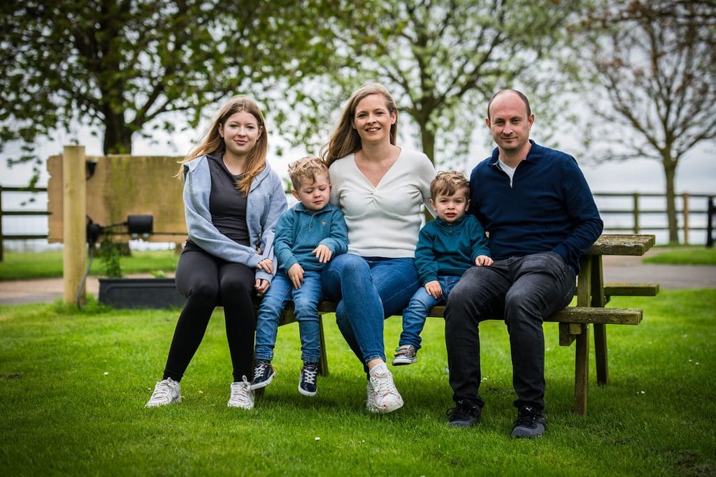 A family of five sitting on a wooden bench in a park, with trees and grass in the background.