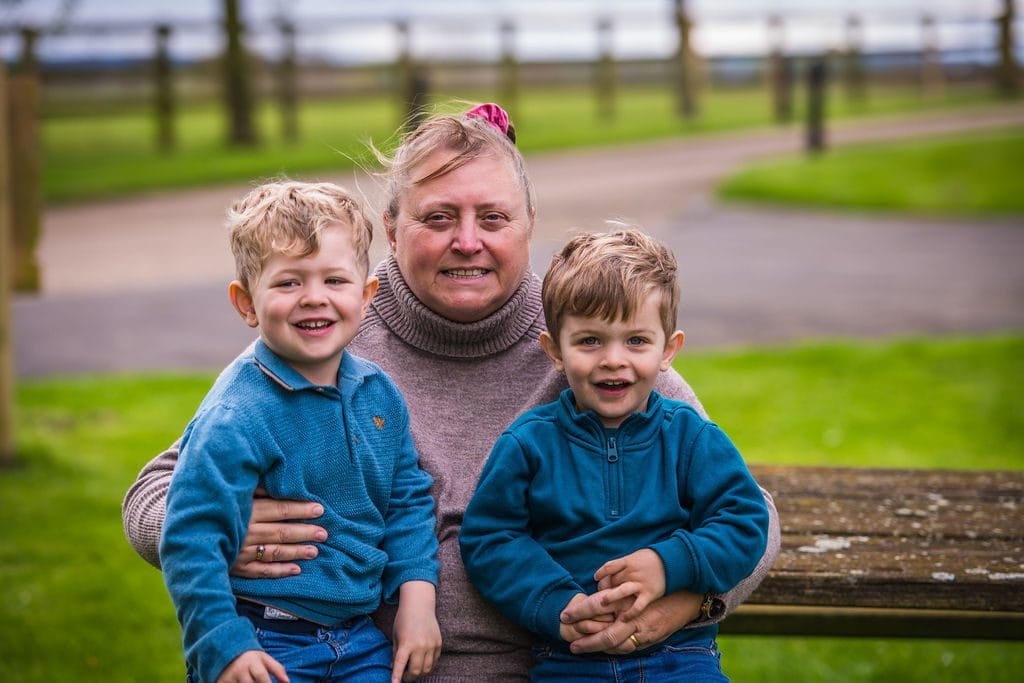 An adult sitting on a bench with two young children, all smiling at the camera, in a park setting.