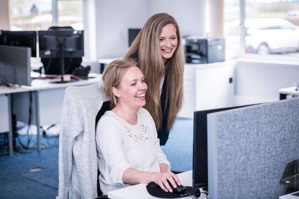 Two women smiling and engaging while working together at a computer in a modern office setting.